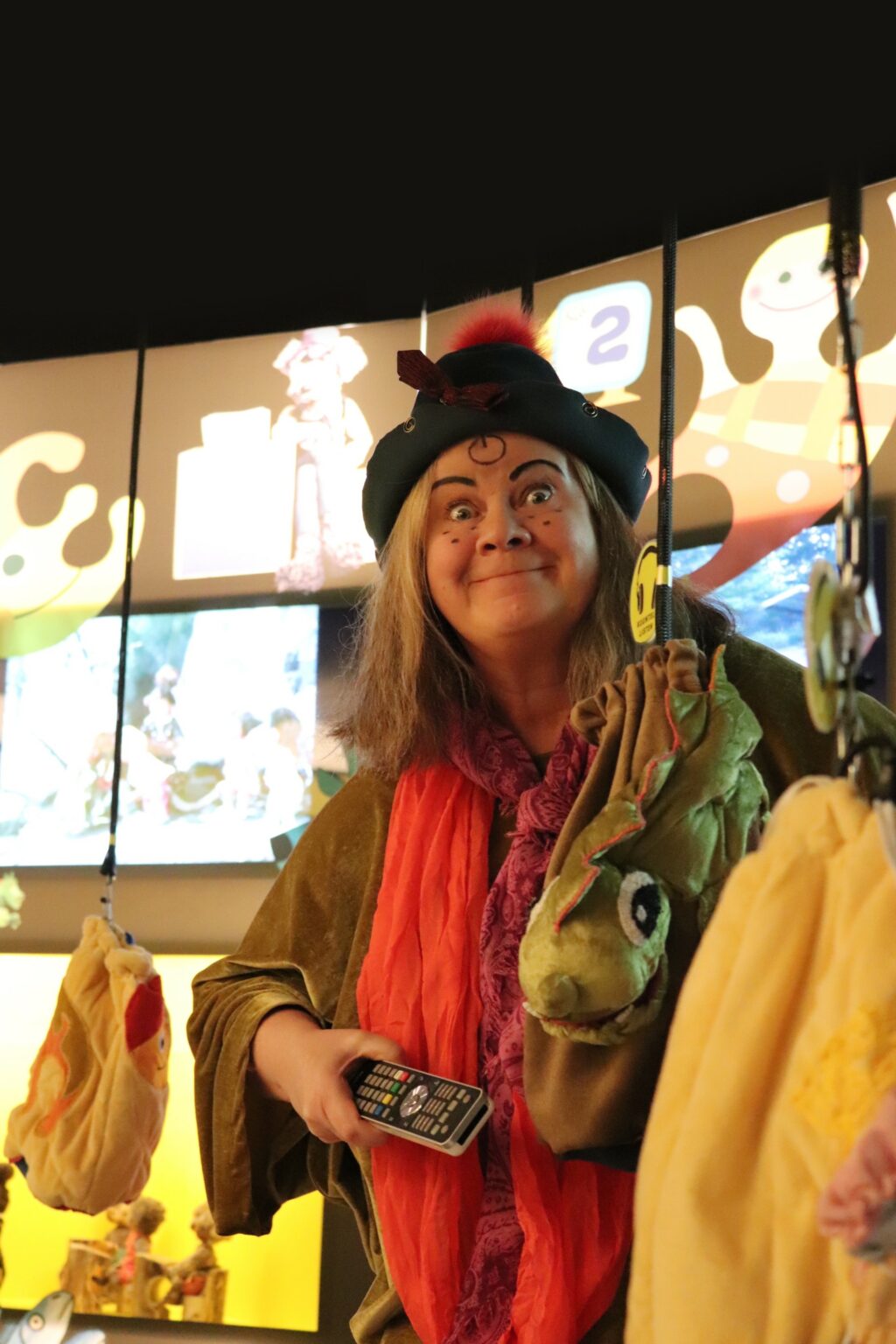 Värikuva oppaasta, jolla on kädessään kaukosäädin ja päällään värikkäät vaatteet. Hänen taustallaan on näyttöjä, joissa näkyy kuvaa erilaisista televisio-ohjelmista.